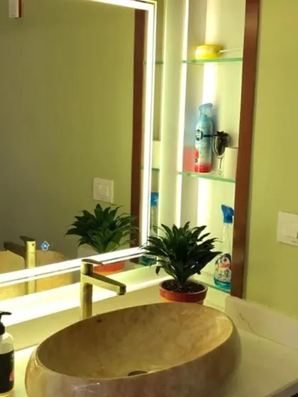 A chic wash area featuring a unique oval-shaped stone vessel sink on a white countertop. The large mirror is framed with built-in vertical lighting and includes a niche with glass shelves for convenient and stylish storage.