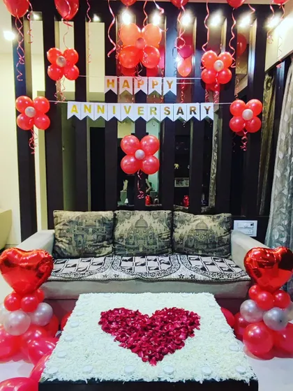 An elegant anniversary decoration in a living room space. The setup features a "Happy Anniversary" banner, red balloon arrangements, and a large heart made from rose petals on the center table.