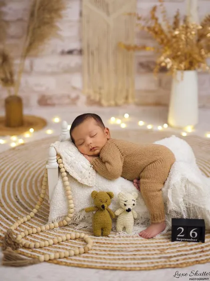 A 26-day-old newborn in a cozy brown knit outfit, sleeping on a miniature bed. The boho-inspired setup with macrame and fairy lights creates a warm and trendy vibe.