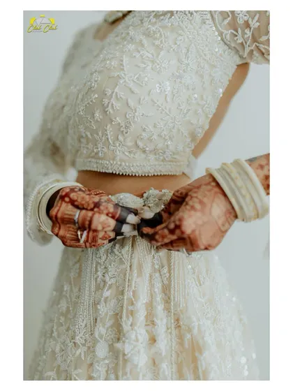 A close-up detail shot of the bride's hands adjusting her lehenga. It highlights the intricate embroidery and the beautiful henna, focusing on the small details that make the bridal look complete.