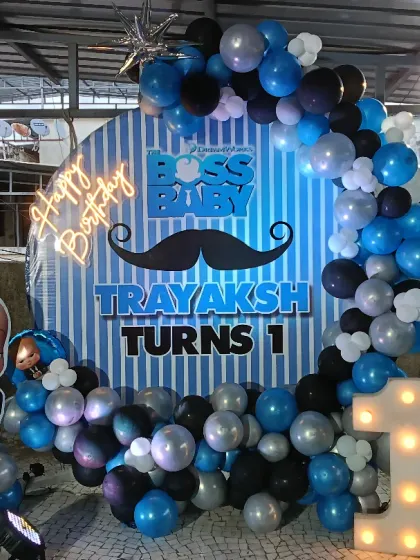 The Boss Baby theme is always fun for a first birthday. This setup includes a circular backdrop, character cutouts, a light-up number '1', and a balloon arch in blue, black, and silver.
