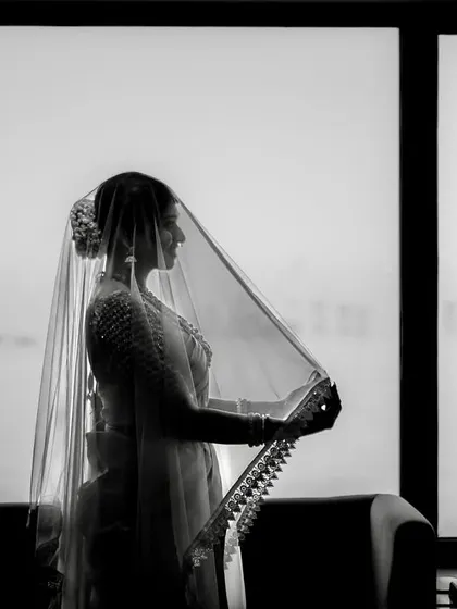 A dramatic black and white silhouette of the bride. She stands against a window, her form veiled, creating a sense of anticipation and timeless beauty.