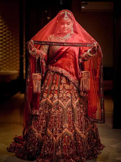 A full-length portrait of a bride in a deep red lehenga, holding her veil. The lighting creates a classic, almost regal look.