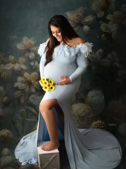 A lovely composition featuring the mom-to-be in a light blue gown against a floral backdrop, holding a small bouquet of sunflowers.