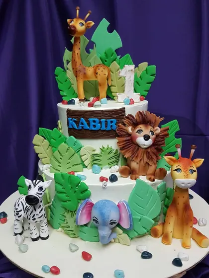 This two-tier jungle animal cake features adorable, handcrafted figures of a lion, zebra, elephant, and giraffe. The lush green leaves and personalized name plaque make it a special cake for a first birthday.