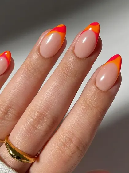 A close-up of a vibrant and fiery double French manicure. The almond-shaped nails have a clean nude base with layered red and orange tips, creating a beautiful sunset effect.