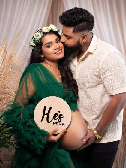 A sweet kiss for the mom-to-be as she holds a "He's Here" sign, a lovely way to do a gender reveal as part of the shoot.