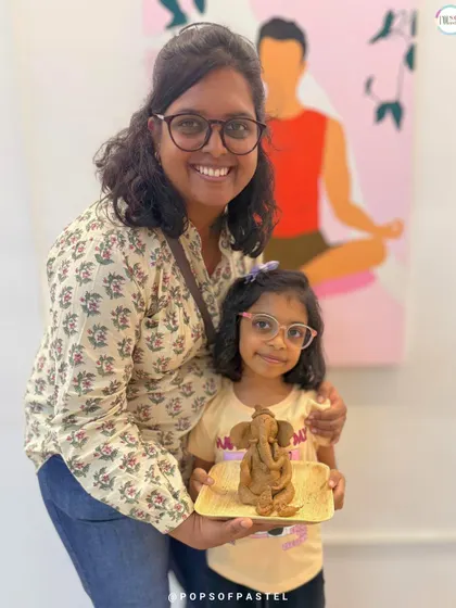 A mother and daughter duo share a happy moment, showcasing the beautiful clay Ganesha they created together in our family-friendly workshop.