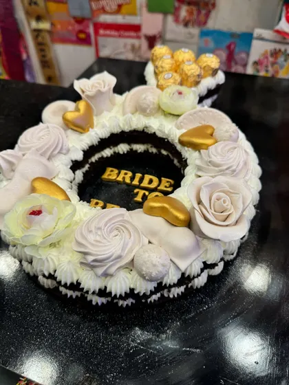A unique ring-shaped "Bride to Be" cake. The design is elegant and creative, with white floral piping and a Ferrero Rocher "diamond," making it a stunning choice for a bridal shower or engagement party.