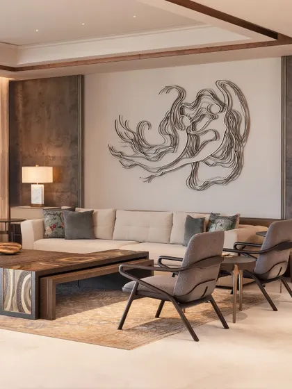This view highlights the harmony between furniture and art. The clean lines of the custom armchairs and coffee table create a grounded setting for the fluid, expressive metal wall sculpture. The entire composition is designed to feel balanced and intentional.