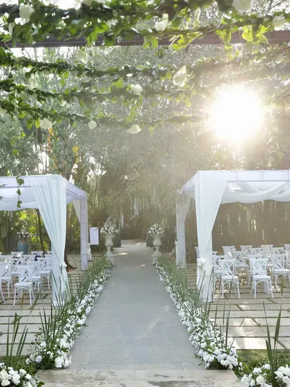 The sun shines down on this elegant outdoor wedding aisle, lined with white chairs and leading to a canopy-covered seating area.