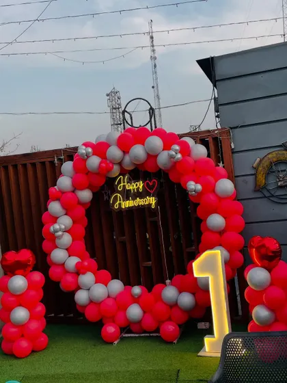 A vibrant first-anniversary party on a rooftop. This setup includes a circular balloon arch in red and silver, a neon sign, and a light-up number '1'.
