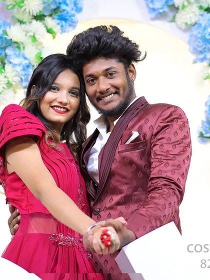 A happy couple poses in our studio, with the woman in a vibrant red tiered gown and the man in a complementary patterned blazer. This shows our coordinated outfits against a floral backdrop.