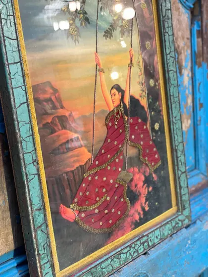 This cloth oleograph, featuring a woman on a swing, is framed with a distressed finish that complements the vintage artwork. The added embroidery gives the piece a unique, three-dimensional appeal.