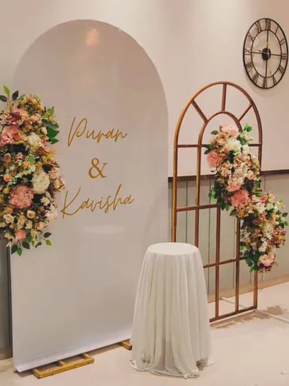 A clean and romantic engagement backdrop. I paired a white arched panel with the couple's names, "Puran & Kavisha," with a rustic window frame accent and soft floral decor.