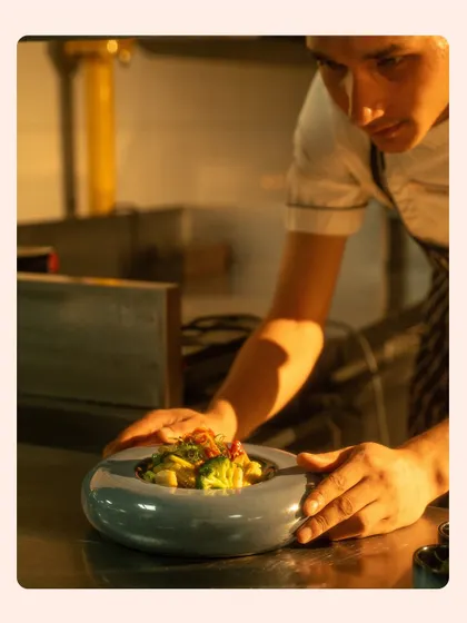 Another angle of a chef plating, with the warm kitchen light catching his hands and the dish. This shot emphasizes the hands-on, careful approach to food preparation.