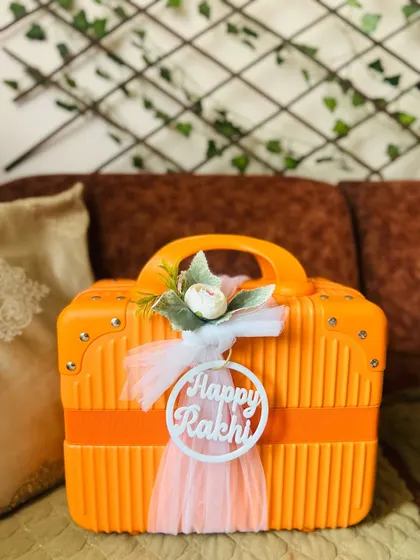 A front view of the decorated Rakhi suitcase hamper. The white "Happy Rakhi" tag contrasts nicely with the bright orange case.