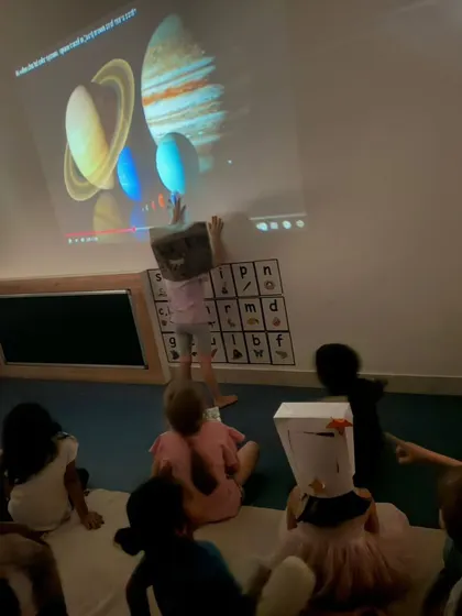 A child points to the projected image of the solar system, fully immersed in the lesson. Using a projector in a darkened room creates a captivating and memorable learning experience.