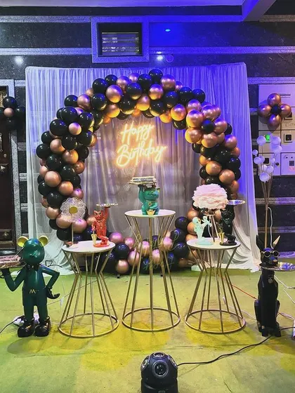 This outdoor setup features a sophisticated black and rose gold balloon arch. The decor is complemented by a neon sign and minimalist plinths, creating a modern and stylish look for an evening celebration.