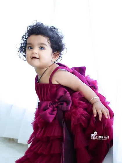 A toddler girl in a beautiful ruffled magenta dress looks up curiously. This studio portrait captures her adorable personality and stylish outfit against a clean, bright background.