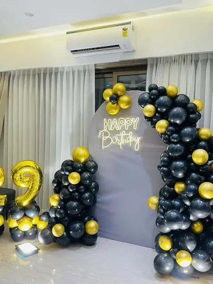 A 29th birthday home decoration with a grey arched backdrop and a neon sign. The black and gold balloon garland and foil numbers complete the modern look.
