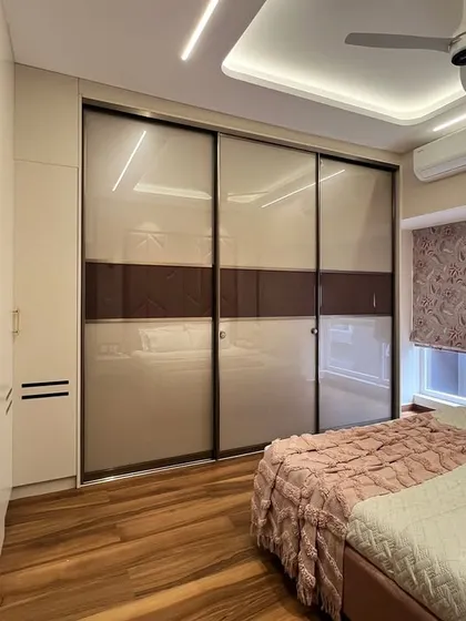 This image shows a sleek, modern wardrobe with sliding doors in a soft beige and brown finish. The design includes open shelving and drawers, providing a mix of concealed and display storage.