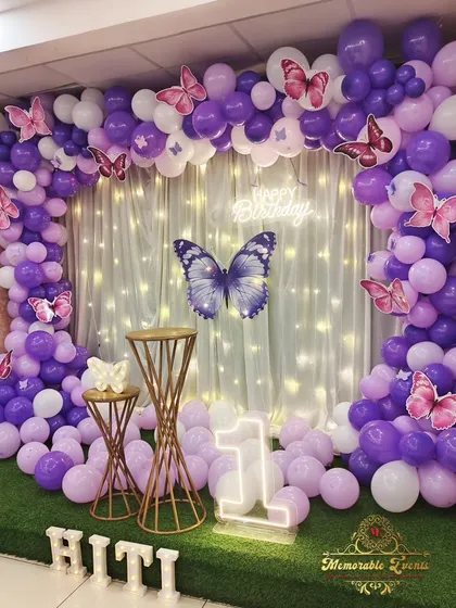 A whimsical butterfly-themed decoration for a first birthday, featuring a purple and white balloon arch against a grass wall, with a neon number '1'.