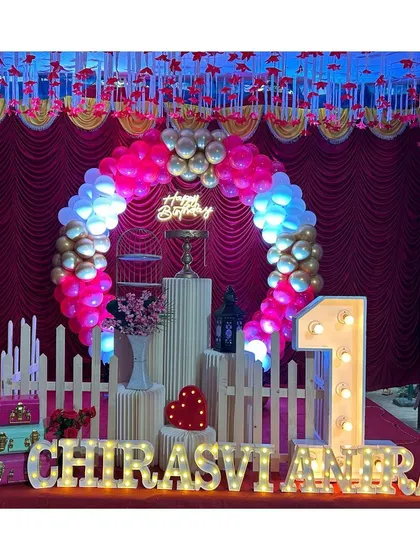 A classic first birthday setup with a circular balloon arch in red, white, and gold, a light-up number '1', and illuminated name letters.