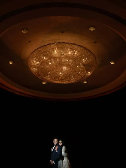 A dramatic, artistic portrait using negative space. The couple stands small under a massive, ornate chandelier, creating a sense of grandeur and intimacy.