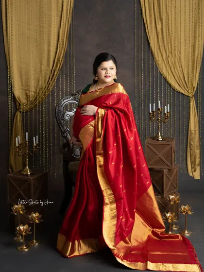 An elegant portrait of an expecting mother in a beautiful red silk saree with a wide gold border. She stands in a traditionally decorated studio with brass lamps and golden drapes.