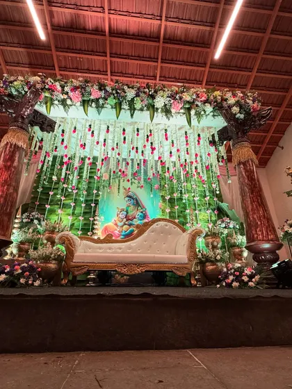 A beautiful baby shower stage featuring a Krishna and Yashoda backdrop, surrounded by hanging floral decorations and traditional pillars.