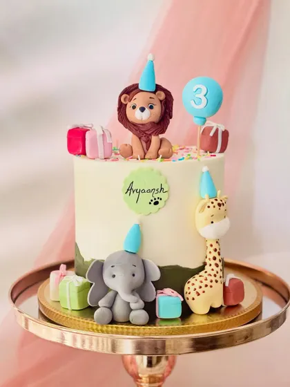 This jungle-themed cake for a third birthday features a lion, elephant, and giraffe in party hats. The design is personalized with the name 'Aryaansh' and a cute paw print.
