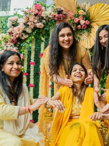 The pure joy of a Haldi ceremony, with friends and family sharing a laugh. The floral backdrop with pastel roses and lush greenery adds a touch of elegance to the vibrant yellow theme.