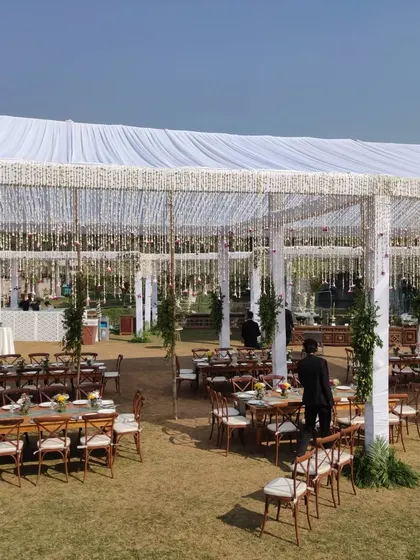 A beautiful and expansive daytime event setup under a large white tent. The decor features elegant guest seating with long wooden tables and hanging floral tassels, perfect for a sophisticated farmhouse lunch or brunch event.