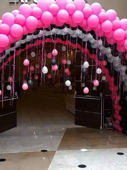 A multi-layered balloon arch designed to create an impressive entrance for a party. The combination of pink, black, and white balloons with hanging elements inside the arch makes for a grand welcome for guests.