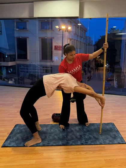 An instructor assists a student in a deep backbend using a stick. This personalized attention ensures safety and helps the student explore their full potential.