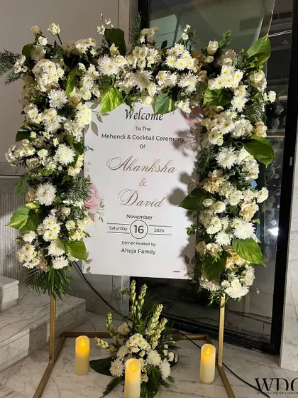 A gorgeous welcome board for a Mehendi and Cocktail ceremony. The sign is framed by an arch of white roses, chrysanthemums, and lush greenery, setting a classy and elegant tone.