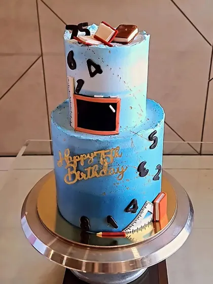 A special cake for a teacher's 75th birthday. This two-tier Swiss Chocolate cake has a blue ombre design and is decorated with fondant books, a chalkboard, and other school-themed items.