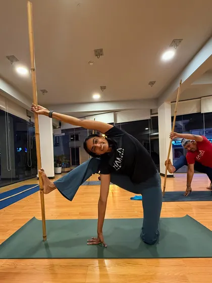 A smiling student holds a challenging side-angle pose with the help of a stick, demonstrating how props can bring both support and joy to the practice.