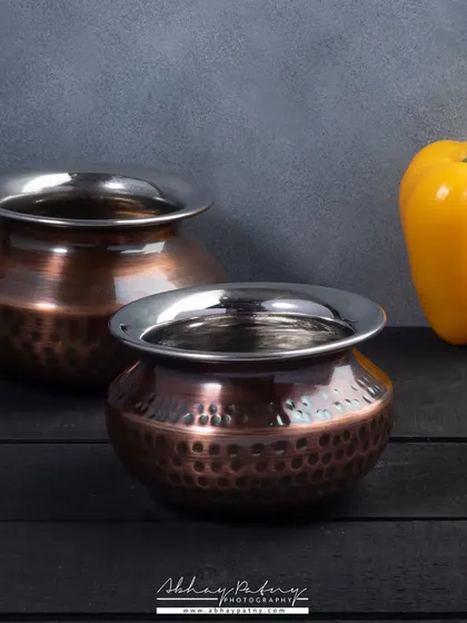 A simple, artistic shot of copper and steel handis. The focus here is on the form and texture of the cookware, using a single prop to add a pop of color and context.
