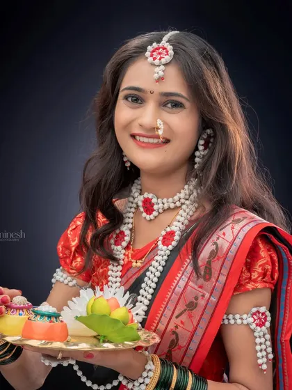 A bright and cheerful portrait of a woman in a red and black saree, holding a decorative puja thali for a Sankranti themed shoot. Her look is completed with traditional sugar jewelry.