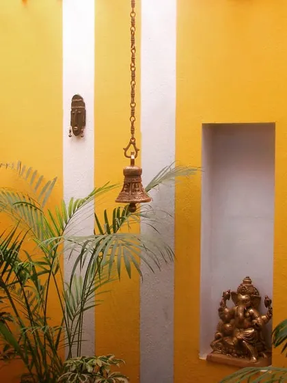 This decorative garden wall serves as an artistic focal point in a residential courtyard. The combination of vibrant yellow paint, traditional brass bells, a niche for a Ganesha statue, and tropical foliage creates a culturally rich and visually stunning feature.