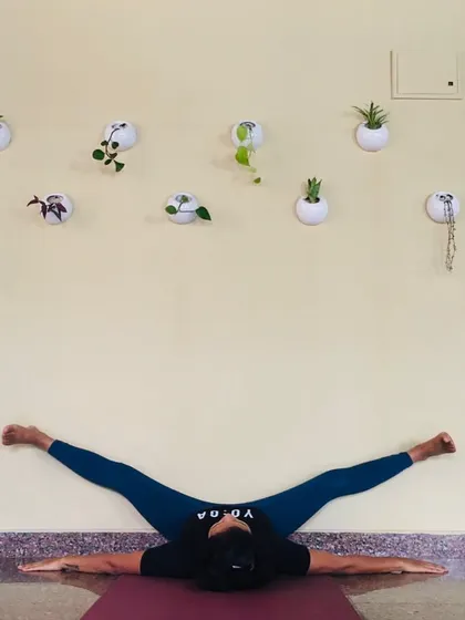 A student rests in a wide legged split against the wall, with our studio plants in the background. Our students are so involved that they even help take care of the plants, making the studio feel like home.