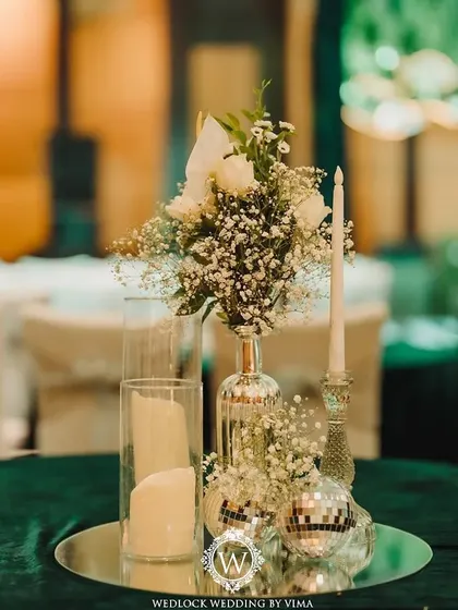 An elegant centerpiece for a Sangeet night, featuring white florals, baby's breath, and candles on a mirrored tray. The mini disco balls add a touch of sparkle and fun, tying into the party theme.