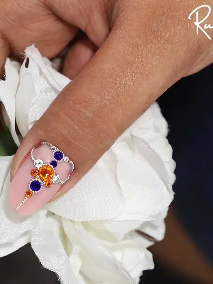 A close-up of a custom jewelry design on a matte pink nail. I used a combination of orange and blue crystals with a delicate silver chain to create this unique, eye-catching piece.