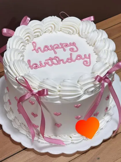 A classic birthday cake with a sweet and simple design. The white buttercream is decorated with piped swirls, pink hearts, and delicate ribbons for a timeless and celebratory look.