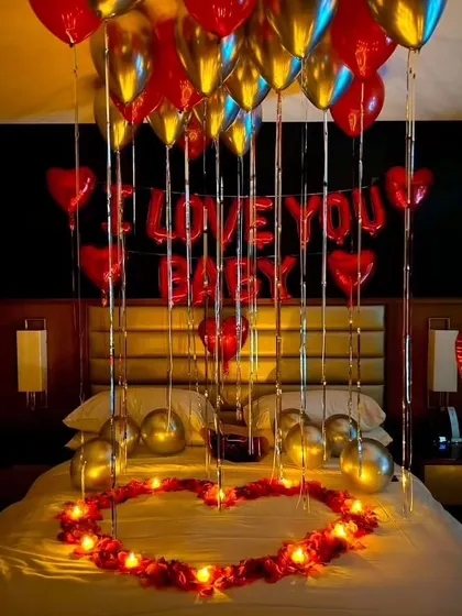 A romantic room setup with red and gold balloons. The decor features 'I Love You Baby' foil balloons, and the bed is decorated with a heart made of rose petals and battery-operated candles.