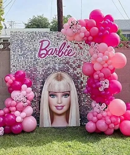 A glamorous outdoor Barbie-themed party. The setup includes a silver shimmer wall, a large Barbie head cutout, and beautiful pink organic balloon garlands.
