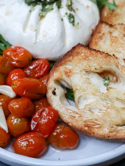 A close-up of our burrata platter, featuring blistered cherry tomatoes, garlic, and perfectly toasted bread. A simple starter that lets the quality of the cheese shine.