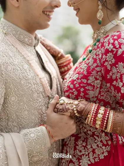 A close-up shot of the couple holding hands, focusing on the bride's intricate red bangles (chooda) and the groom's classic sherwani. A beautiful detail of their wedding attire.